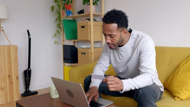 Young adult man paying online with credit card on laptop computer. African american guy shopping on internet making remote payment from home.