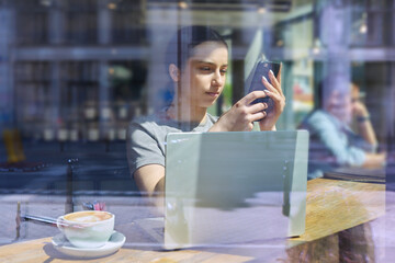 Young female inside cafe with cup of coffee by laptop smartphone looking through window