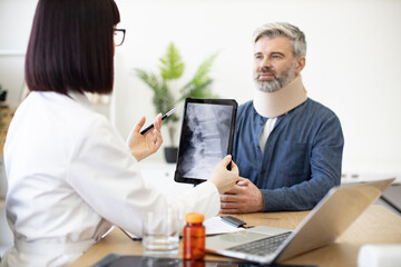 Back view of female doctor holding digital tablet while sitting in front of smiling caucasian man in cervical collar. Health professional consulting mature patient about neck injury using CT scan.