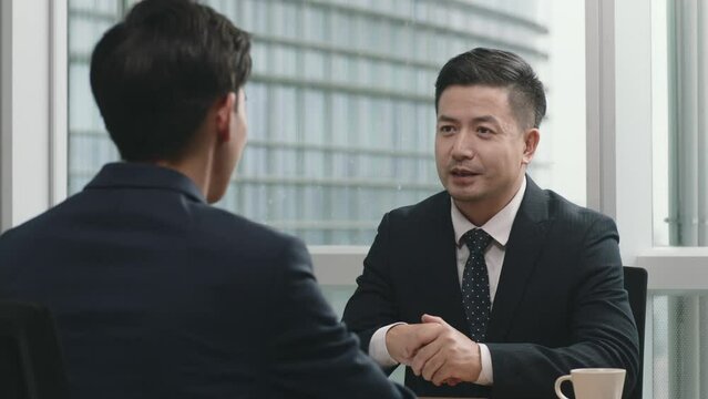 two asian corporate executives sitting at desk having a business discussion shaking hands in modern office