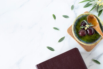 Pure olive oil with olives and wooden spoon, closed Bible Book, and green branch on white. Symbol...