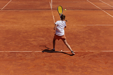 A young girl showing professional tennis skills in a competitive match on a sunny day, surrounded by the modern aesthetics of a tennis court.