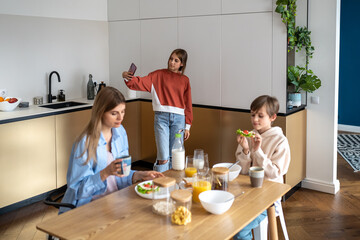 Obraz na płótnie Canvas Family morning, teen daughter making selfie for social media on smartphone, mom son having breakfast sitting at table on kitchen. Motherhood, rising children, parenting, life with teenagers concept.