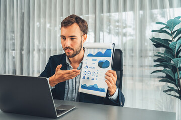 Professional businessman in black suit present financial data or BI paper clipboard to laptop camera during online presentation meeting. Remote work with virtual meeting presentation in office. Entity