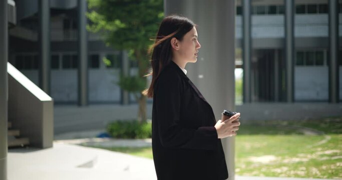 Side view shot, Footage of Young smiling elegant Asian busy business woman in a suit holding a cup of coffee and walking in front of a modern business building. Business and people concepts.