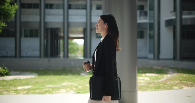 Side view shot, Footage of Young smiling elegant Asian busy business woman in a suit holding a cup of coffee and walking in front of a modern business building. Business and people concepts.