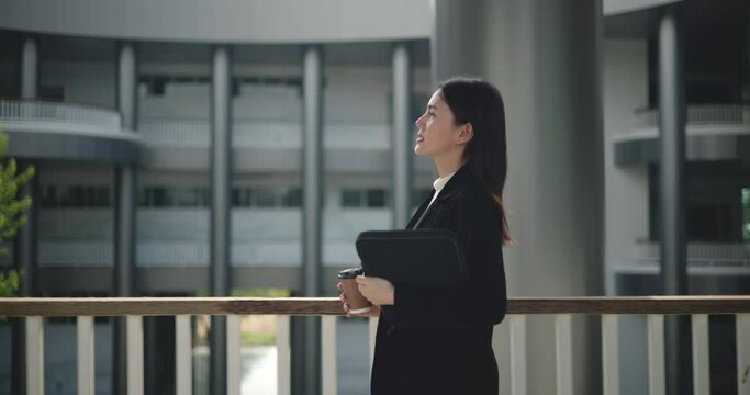 Side view shot, Footage of Happy young Asian businesswoman in suit holding a laptop and cup while walking in a modern business building. Business and people concepts.