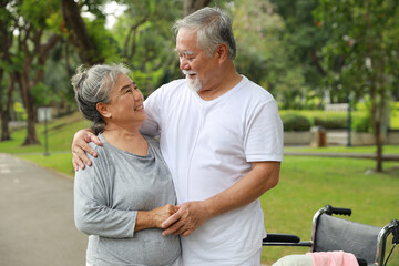 Obraz premium Asian senior woman or caregiver helping senior man walk with wheelchair at park outdoor. Elderly wife taking good help care and support of elder husband patient outside the retirement house.