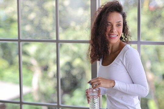 Charming African American holding and drinking bottle of water drink clean clear mineral aqua for healthy and body refreshment. Happy female standing in living room at home