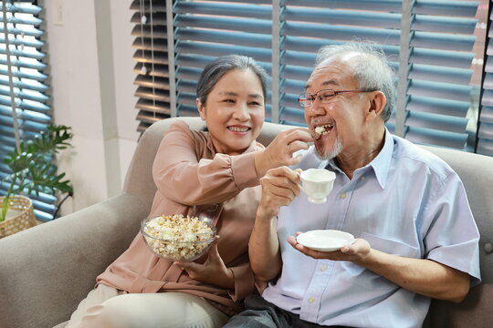 Happy Smiling Asian Senior Man And Woman Sitting On Sofa And Eating Popcorn While Having Fun With Movie Rest Indoor At Home Living Room. Couple Elder Husband And Wife Embrace Are Happy While Watch TV