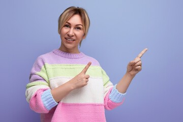 portrait of a pleasant smiling blond young woman in a casual outfit showing a wall with a finger on a purple background with copy space