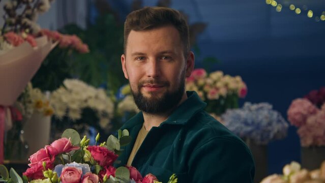 Professional male florist keeps in hands beautiful bouquet, smiles and looks at camera. Vases with fresh flowers stand at background. Concept of retail floral business and entrepreneurship. Portrait.