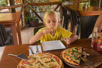 An american curly blonde boy eating a pizza in italian restaurant. Portrait of child with long...