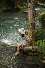 Cute curly woman dressed in a dress and a straw hat sits near the river in the tropical jungle. Lonely girl tourist in the forest. Concept - travel around the world.