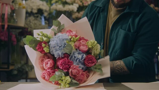 Male florist wraps beautiful bouquet, uses wrapping paper. Colleague with vase of flowers walks at background. Process of working in flower shop. Floral business and entrepreneurship concept. Close up