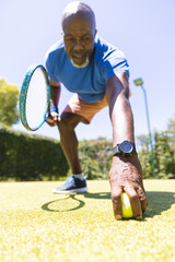 Senior african american man bending down to pick up tennis ball on sunny grass court