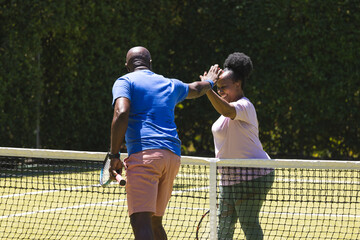 Happy senior african american couple high fiving over net on sunny grass tennis court