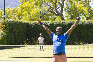 Happy senior african american couple with tennis rackets, man celebrating on sunny tennis court
