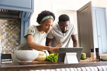 Tuinposter Focused african american couple preparing meal together using tablet in kitchen © wavebreak3
