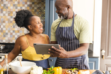 Happy senior african american couple preparing meal together, using tablet in kitchen
