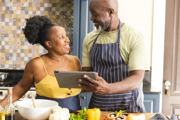 Tuinposter Happy senior african american couple preparing meal together, using tablet in kitchen © wavebreak3
