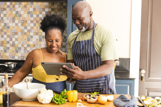 Happy senior african american couple preparing meal together, using tablet in kitchen
