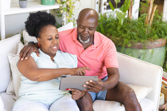Happy Senior African American Couple On Couch Using Tablet And Embracing In Sunny Living Room