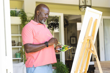 Happy senior african american man holding paint palette painting canvas on easel at home - Powered by Adobe