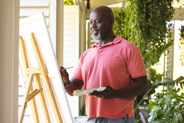 Happy senior african american man holding paint palette painting canvas on easel at home