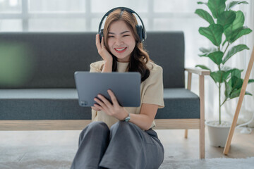 Happy young woman wear headphone with using tablet in living room on sofa. Young happy woman lie playing smart tablet at home. Beautiful young asian women using tablet computer.