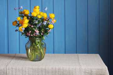 Yellow globe flowers and Forget-me-nots in a glass jar on a blue background. Cute bouquet of wildflowers.