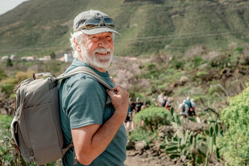 Happy caucasian senior man with hat and backpack looks at camera walking the trail on a mountain...