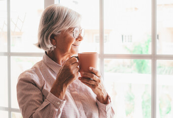 Portrait of senior caucasian woman at home looking out the window holding a cup of tea, pensive elderly woman drinking a coffee