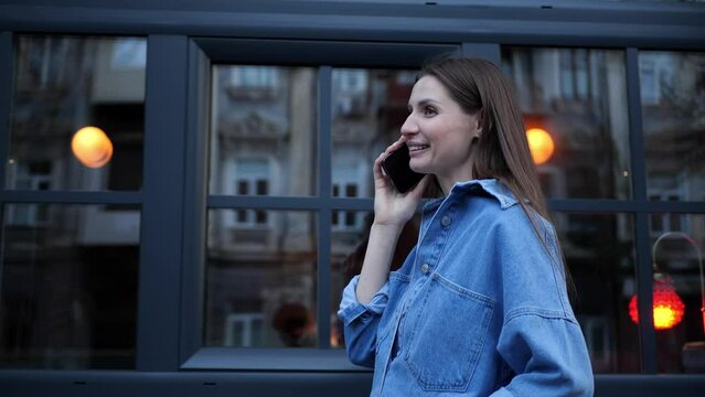 Beautiful young woman talking on phone while walking in city, low angle view. Camera moving left, slow motion