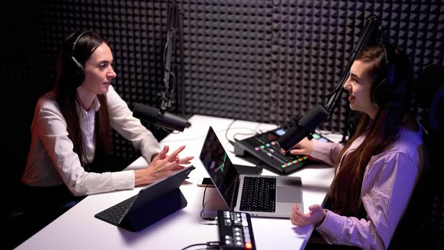 Side angle from above view of young woman podcaster speaking to her female guest ito microphone, both wearing headphones in a modern dark video studio 