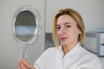 Pretty beautiful girl with natural beauty, holding a mirror in hand, dressed in a white robe, looks at herself after beauty procedures, such as carbon peeling and skin rejuvenation, in beauty clinic.