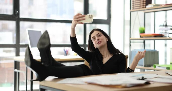 Relaxed manager takes selfie on smartphone at workplace. Woman putting feet on table and posing for photo in office
