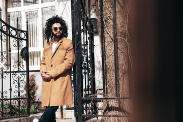 Handsome smiling hipster model.  Unshaven Arabian man dressed in brown coat clothes. Fashion male with long curly hairstyle posing outdoors in street. Cheerful and happy. In sunglasses. At sunny day