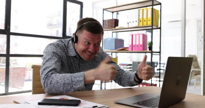Smiling businessman in headphones looking at laptop screen, watching good webinar showing thumbs up. Mood swings consulting client or learning languages remotely showing gesture fak participating in