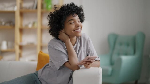 Smiling falling in love african american girl dreaming enjoying reading message in smartphone sitting on sofa at home. Online dating, romantic relationship, cute communication with boyfriend concept.