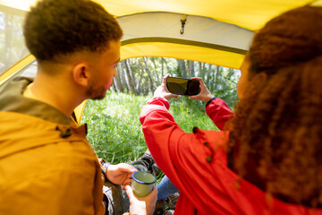 Happy biracial couple sitting in tent and using smartphone in forest, unaltered