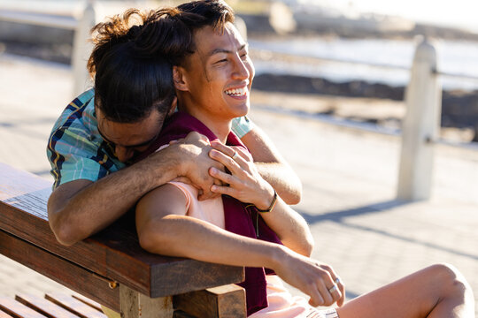 Happy biracial gay male couple sitting on bench and embracing on promenade by the sea