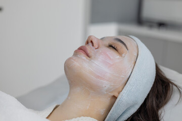 Stunning brunette woman with a white cream mask on her face reclines in a beauty and wellness clinic. Girl luxuriates in skin rejuvenation and care treatments at a spa salon, light smile on face.