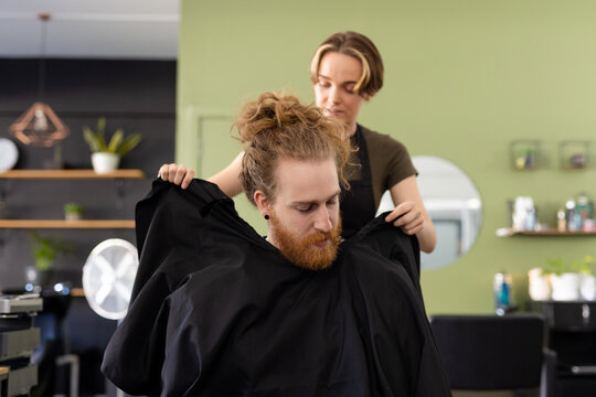 Caucasian female barber putting cape on male customer at barbershop