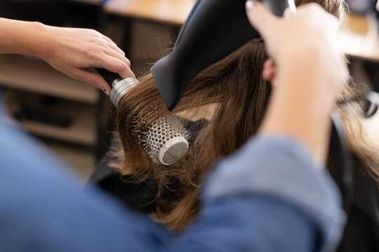 Hands of caucasian female hairdresser styling hair of customer with hairdryer and brush at salon