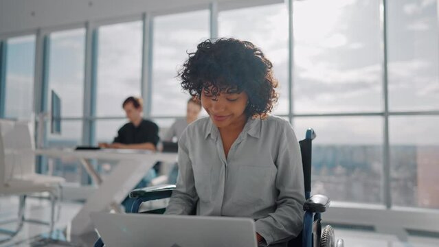 African american woman in wheelchair working on project on laptop in office looking at screen and smiling received nice message from colleagues. Man and woman working sitting at desk on background.