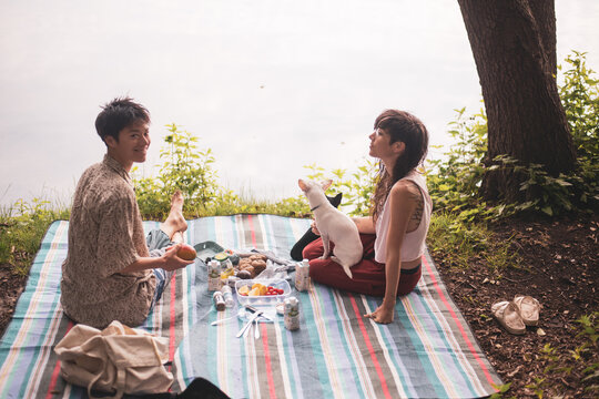 couple sits smiling with dogs on picnic rug by natural lake in europe