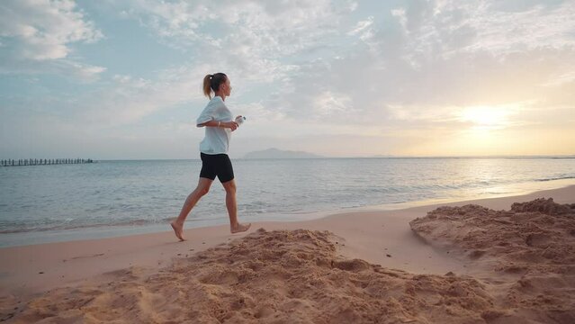 Barefooted woman jogging along sandy sea ocean beach holding bottle of water at sunset. Outdoors training, running, wellness sport training, workout of female. Active lifestyle, body care concept.