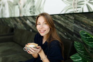 Portrait of joyful young woman enjoying a cup of coffee at home. Smiling pretty girl drinking hot drink. Excited woman wearing blue dress in green interior, smelling cappuccino
