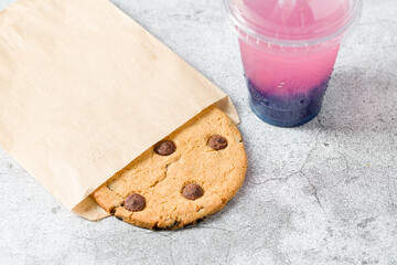 Chocolate chip cookie wrapped in paper bag with drink next to it on stone table